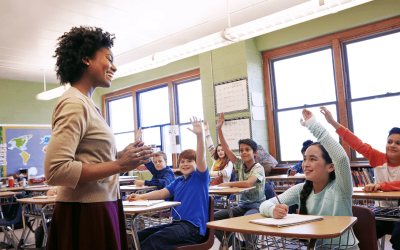School teacher standing at the front of the classroom asking the children a question.